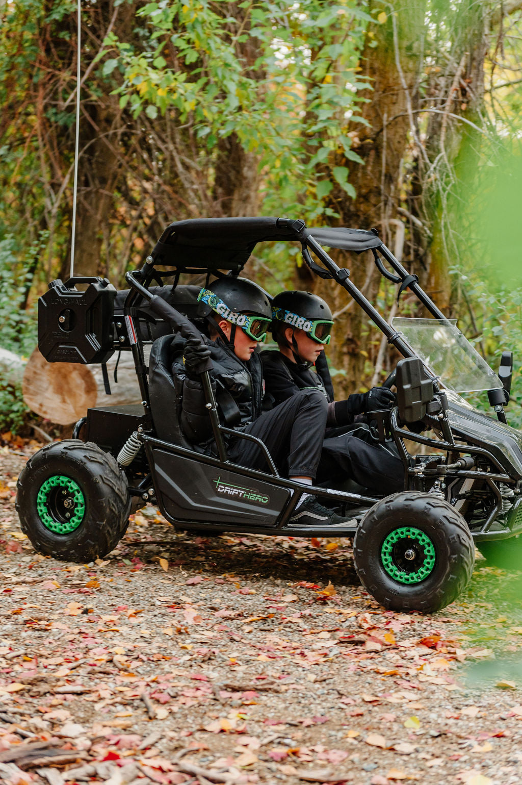 Two people in a Drift Hero USA 2 Seater Electric Go-kart with green wheels on a forest path.