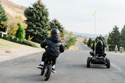 Two children riding a bike and a Drift Hero USA 1 Seater Electric Go-kart on a road with trees and mountains in the background.