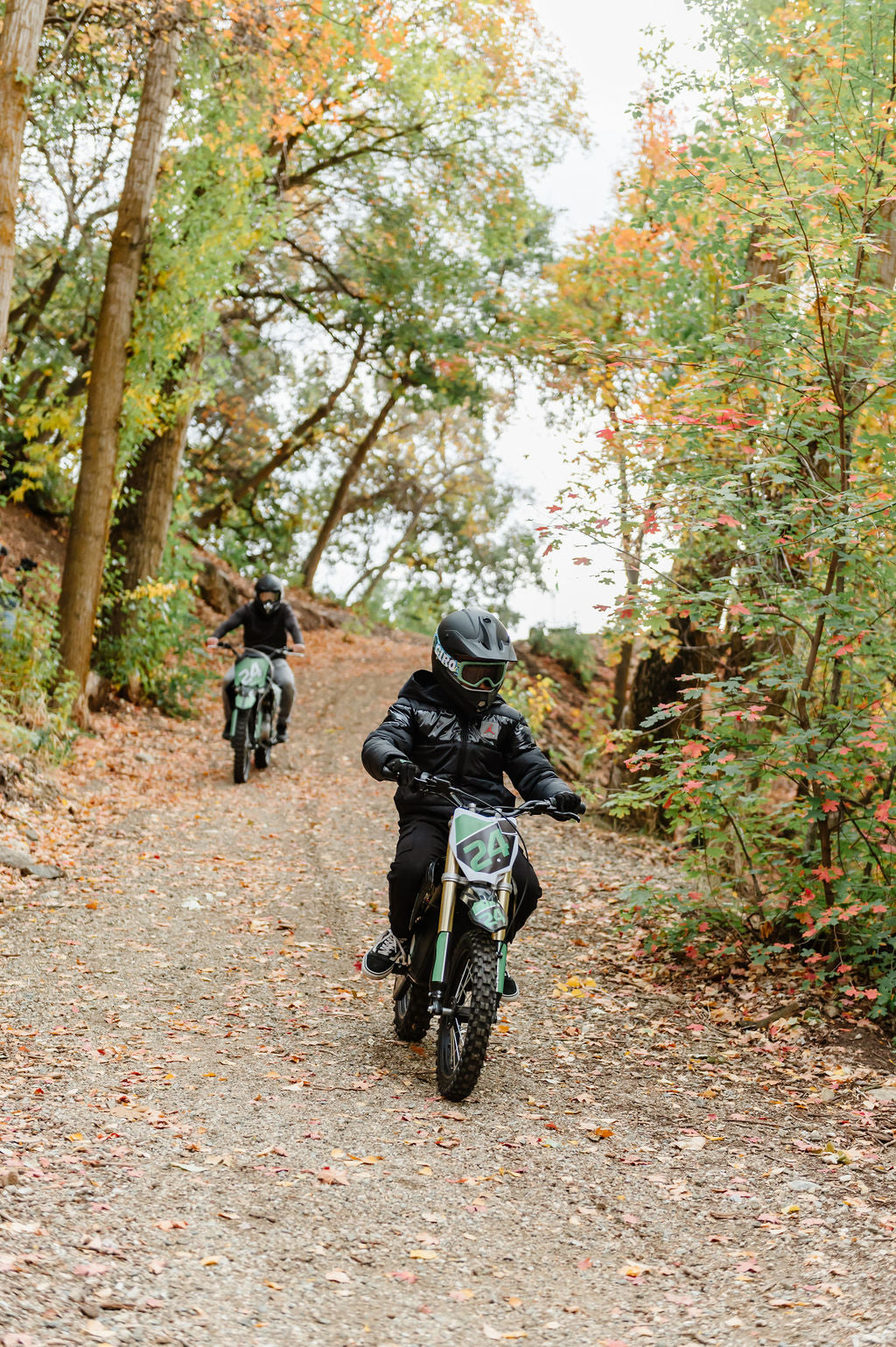 Two people riding Drift Hero USA 1600W Electric Dirt Bike on a forest trail with autumn foliage.