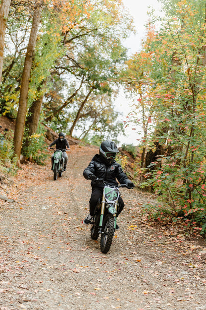 Two people riding Drift Hero USA 1600W Electric Dirt Bike on a forest trail with autumn foliage.