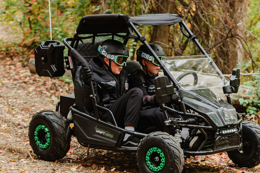 Two children in a Drift Hero USA 2 Seater Electric Go-kart with green wheels driving on a forest trail