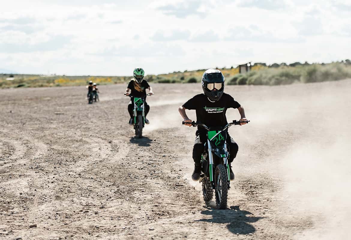Three kids riding Drift Hero USA 800W Electric dirt bike on a dusty track with a clear sky.