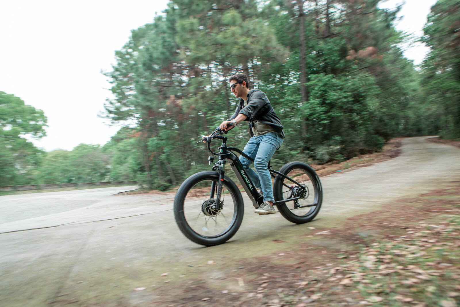 Male rider cycling on a Eunorau FAT AWD 2.0 black ebike outdoors on a forest road with trees in the background