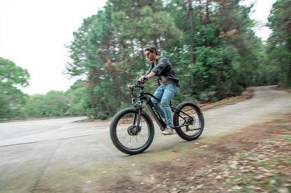 Male rider cycling on a Eunorau FAT AWD 2.0 black ebike outdoors on a forest road with trees in the background