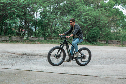 A male rider cycling with a Eunorau FAT AWD 2.0 black ebike on a forest trail with trees in the background