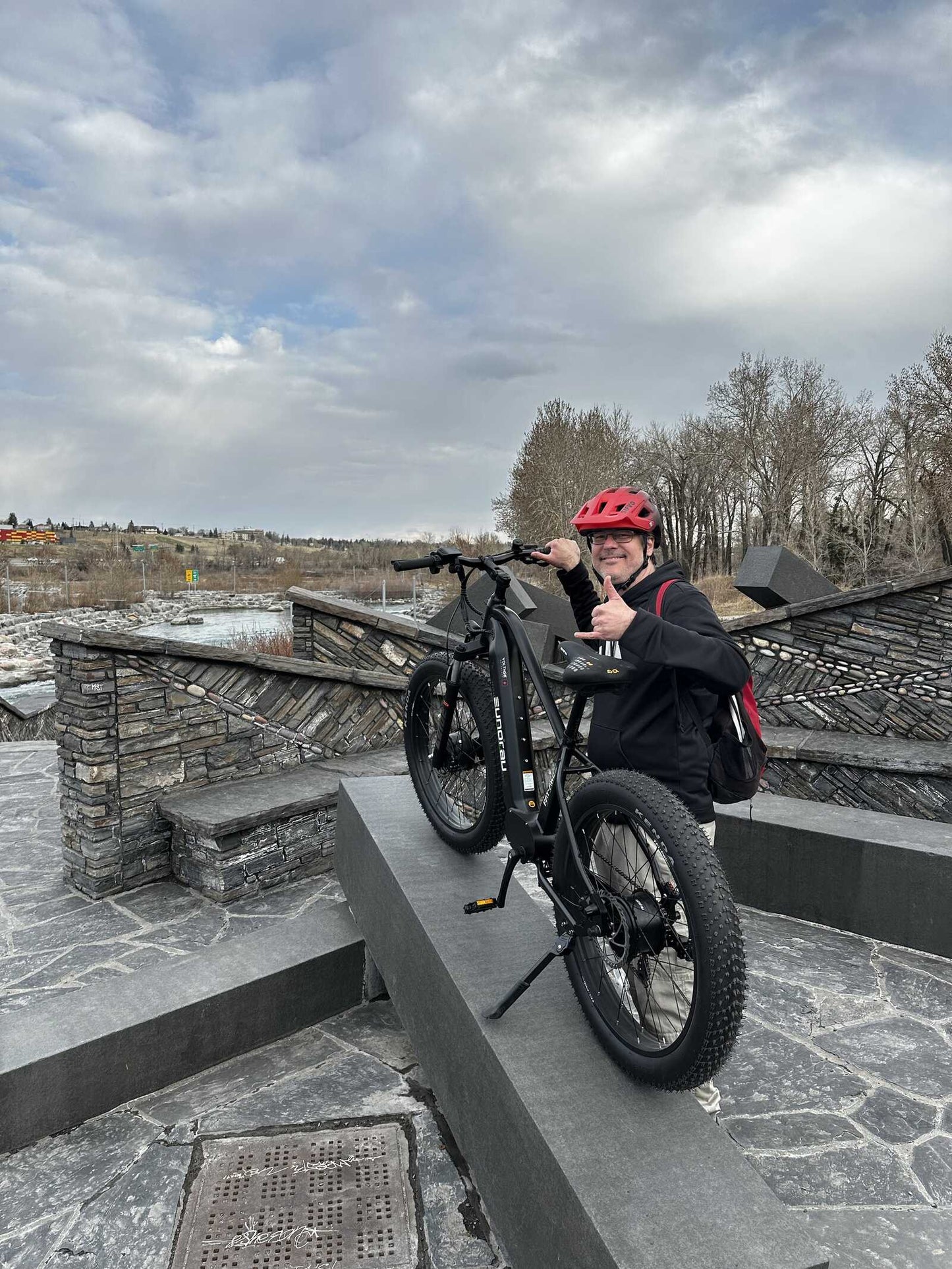 Eunorau FAT AWD 2.0 electric bike, person riding the bike outdoors while wearing a red helmet, posed on a stone structure.
