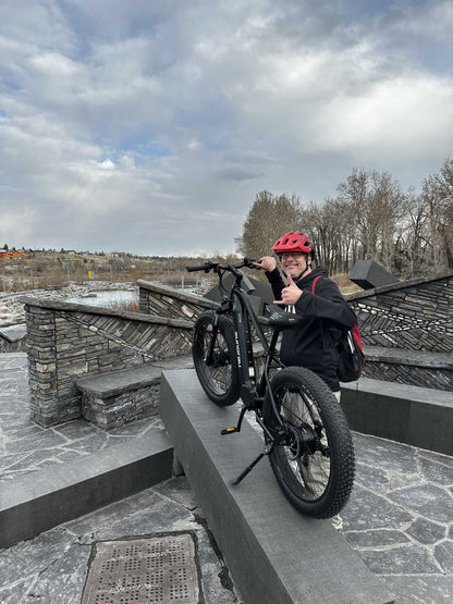 Eunorau FAT AWD 2.0 electric bike, person riding the bike outdoors while wearing a red helmet, posed on a stone structure.

