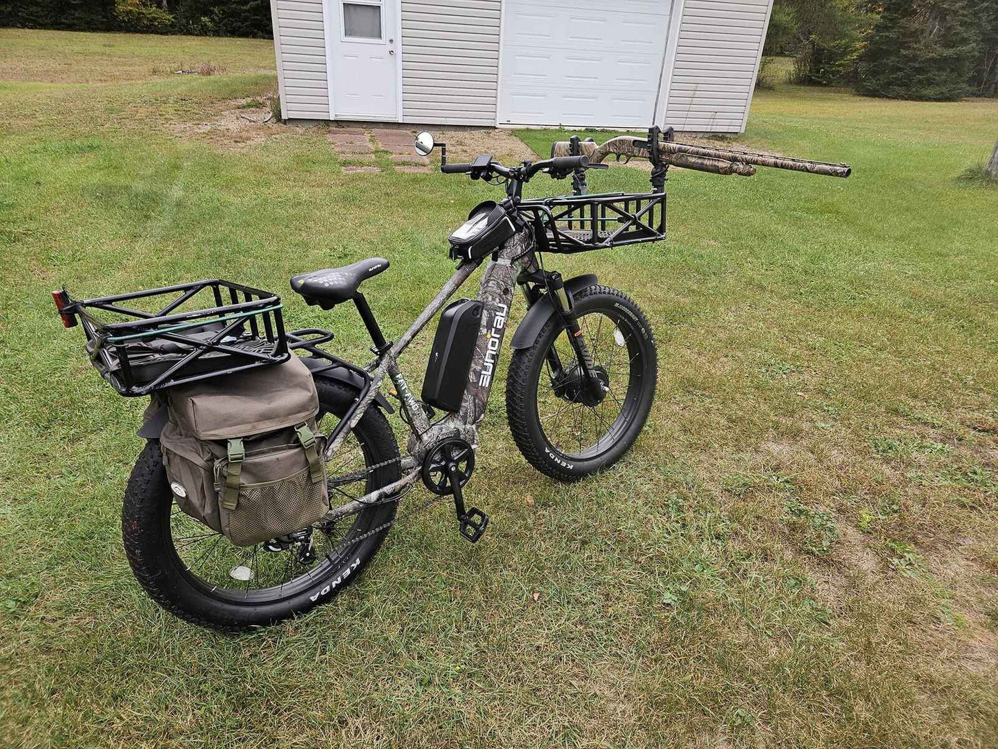 Eunorau FAT AWD 2.0 electric bike in camo finish, left side view on grass with front and rear racks, saddlebag, and mounted hunting rifle.
