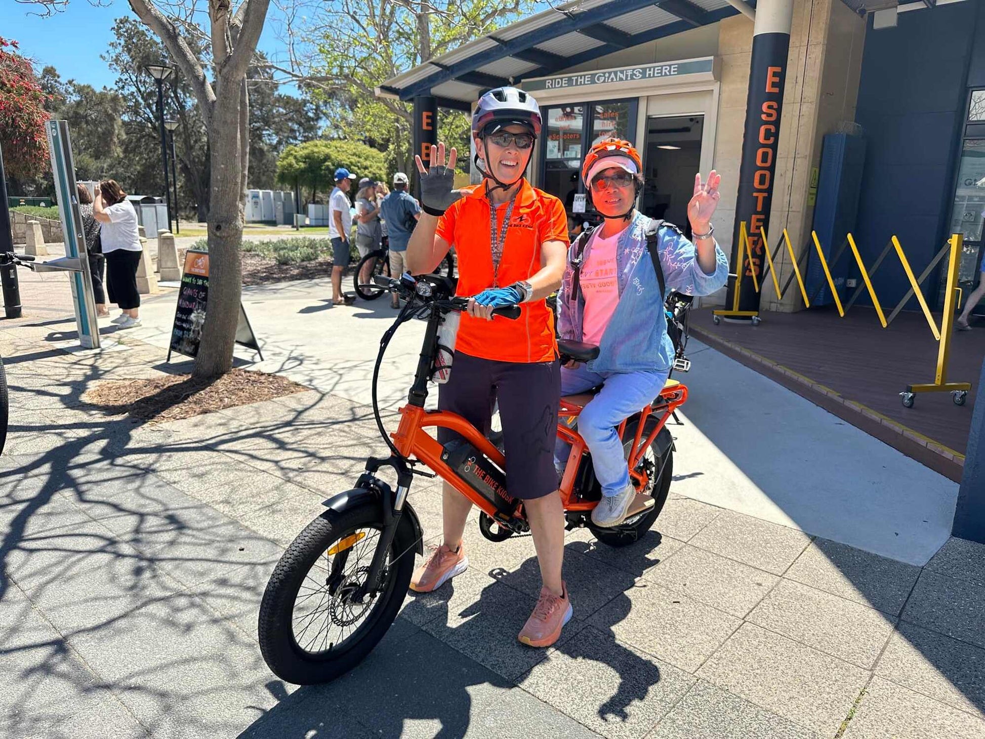 Two people on an Eunorau G30 Cargo electric bike in a park-like setting with a building in the background.