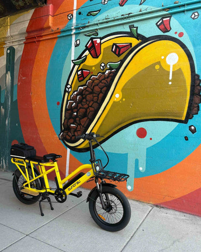 Yellow Eunorau G30 Cargo electric bike parked in front of a colorful mural with a large taco illustration.