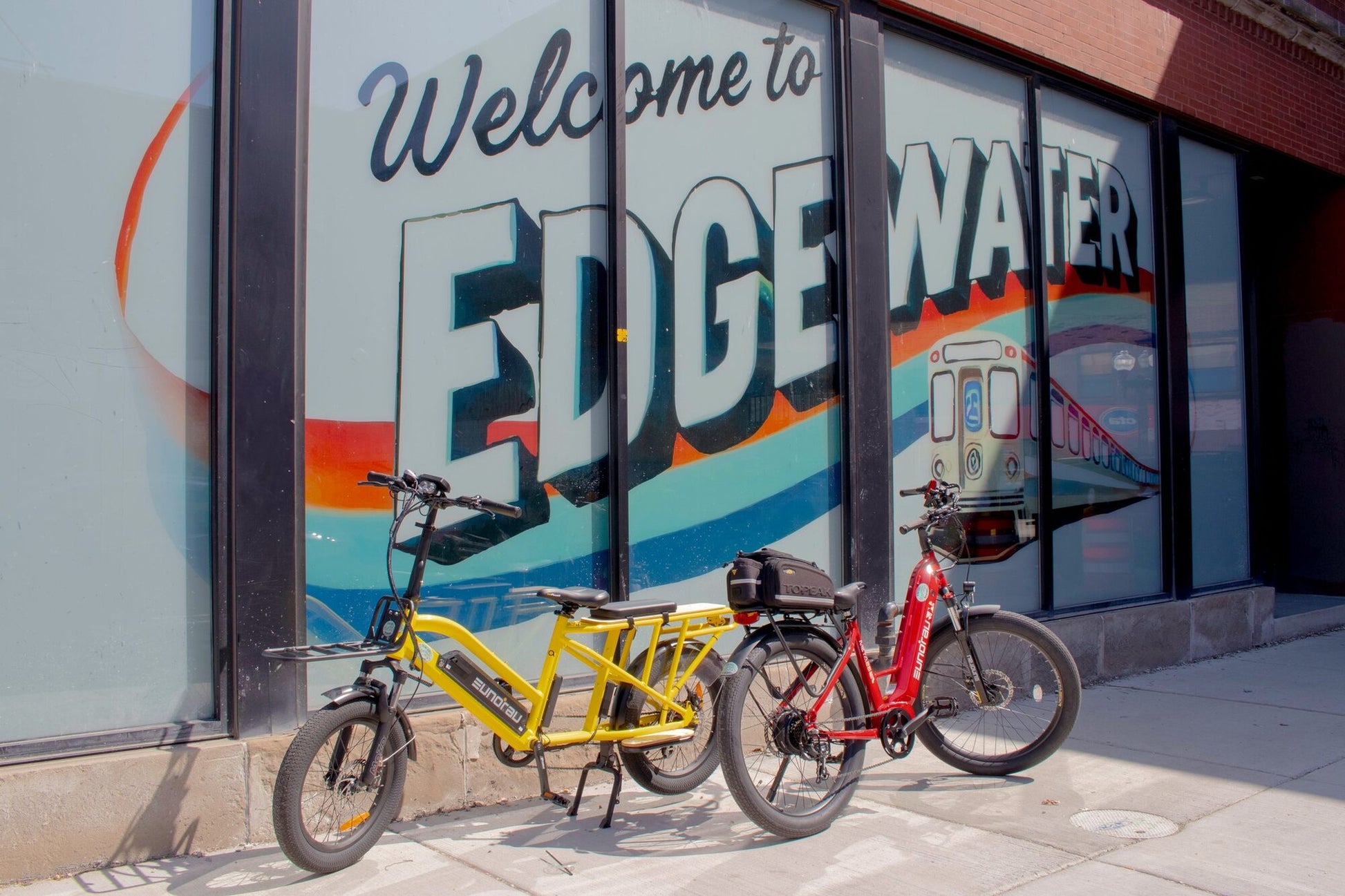 Two Eunorau G30 Cargo electric bicycles, one yellow and one red, parked in front of a store with 'Welcome to Edge Water' sign.