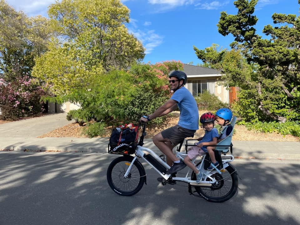 Man riding a Eunorau Max-Cargo 14AH  with two children in a bike trailer on a sunny day.