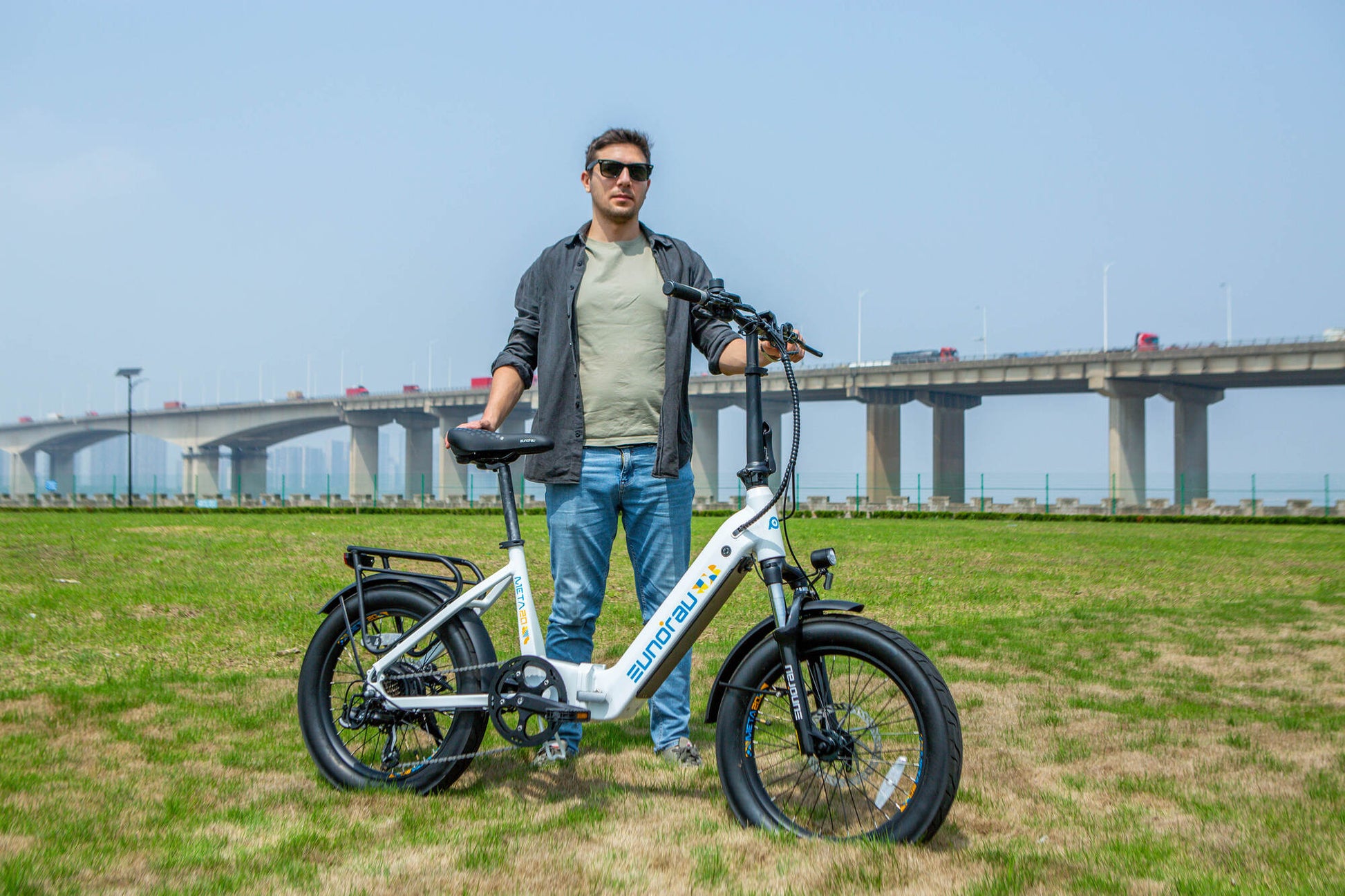 Eunorau Meta 20 Foldable electric bicycle on grass with man holding the bike with a bridge in the background