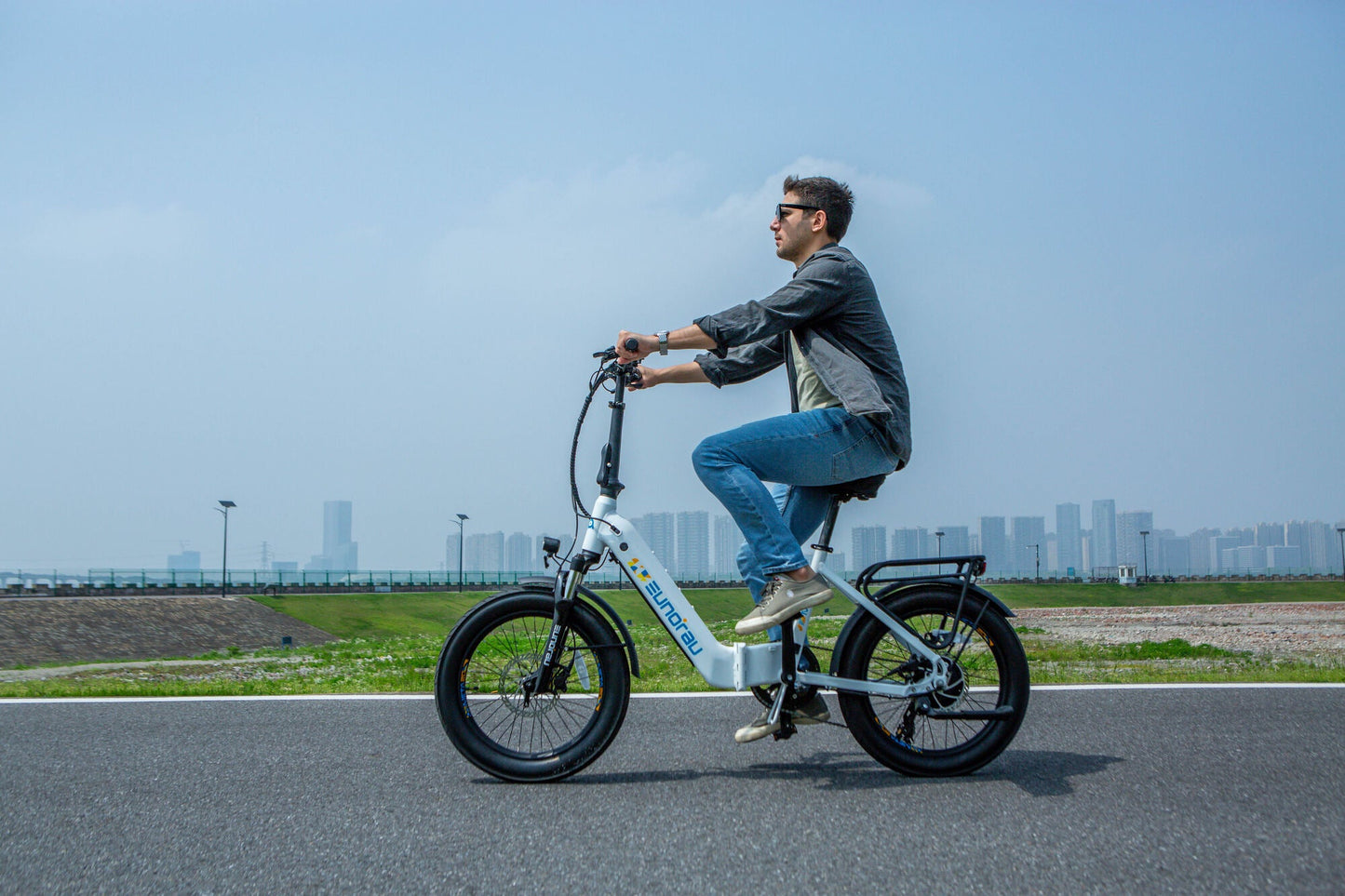 Man riding EUNORAU META Foldable electric bike with 20-inch wheels on urban road with city skyline background