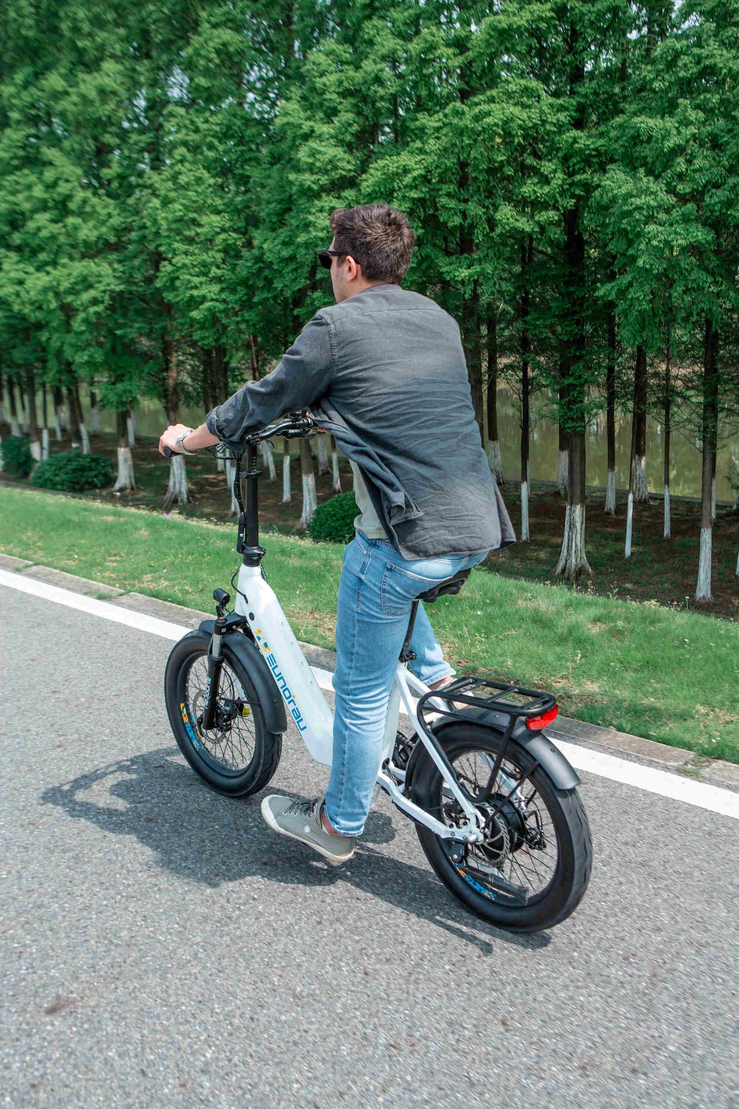 Man riding white EUNORAU META Foldable electric bike with 20-inch wheels on a road surrounded by trees