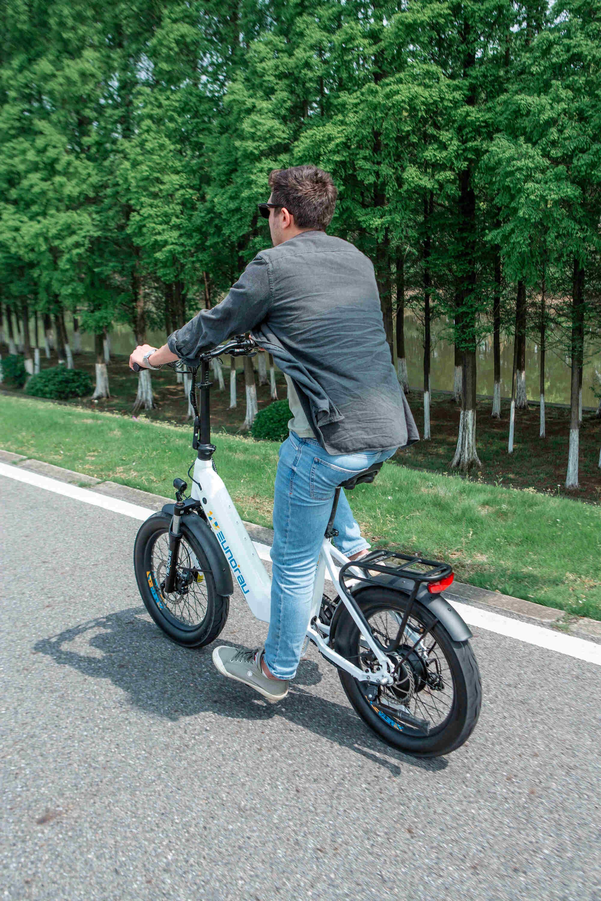 Man riding white EUNORAU META Foldable electric bike with 20-inch wheels on a road surrounded by trees