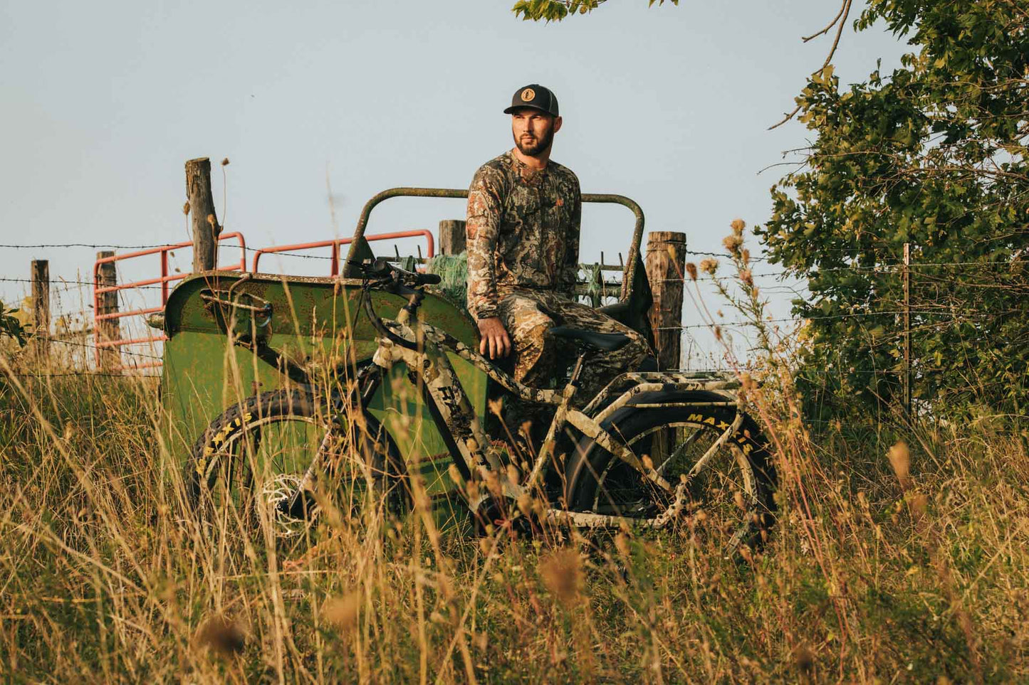 Man in camouflage sitting on a Rambo Bikes Venom 2.0 Electric Bike in a field with trees and a fence in the background.