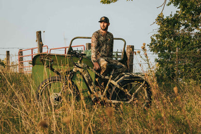 Man in camouflage sitting on a Rambo Bikes Venom 2.0 Electric Bike in a field with trees and a fence in the background.