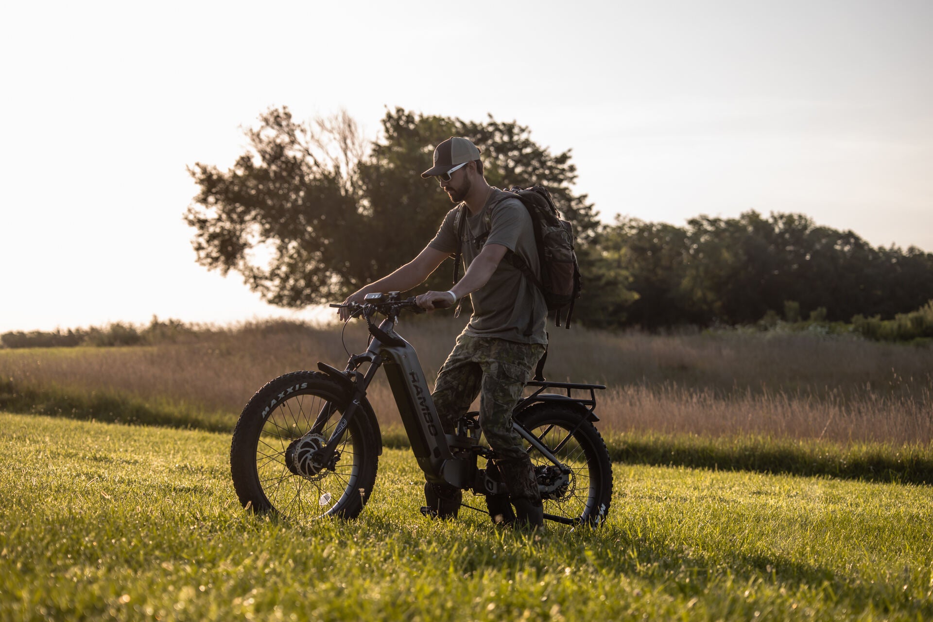 Man with a backpack riding an Rambo Bike Hellcat 2.0 AWD Electric Bike in a field with trees in the background