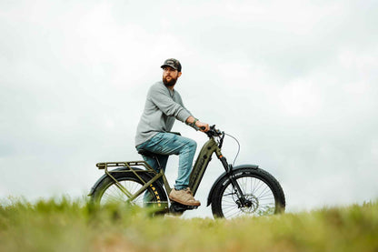 Man riding an Rambo Bikes REBEL 2.0 Electric Bike in a grassy field with a cloudy sky.