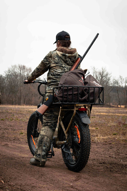 Person on a Rambo Bikes SAVAGE 2.0 Electric Bike with a large bag and rifle, riding through a field.