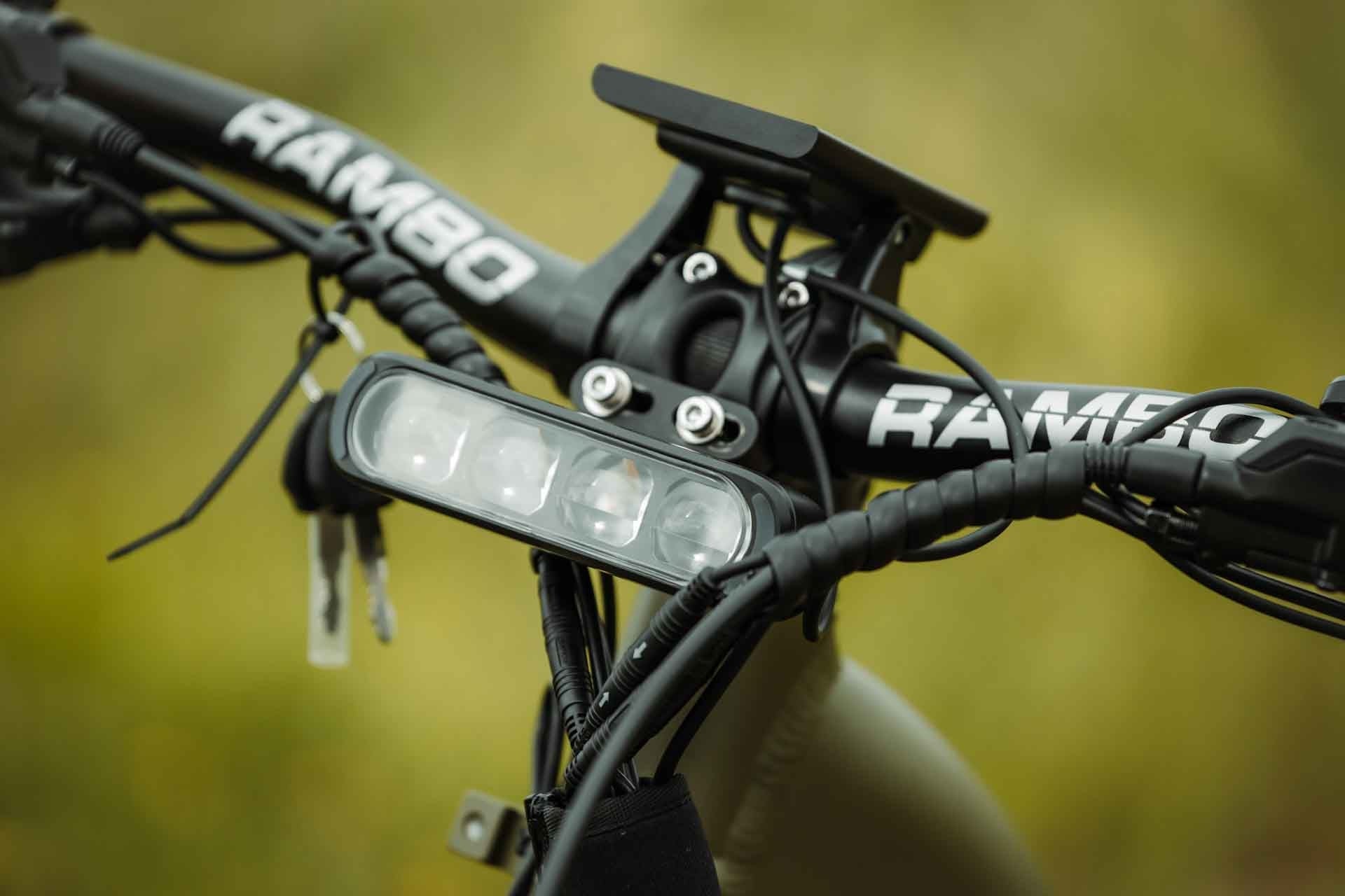 Rambo Bikes REBEL 2.0 Electric Bike handlebar with 'RAMBO' branding and an LED light on a blurred natural background