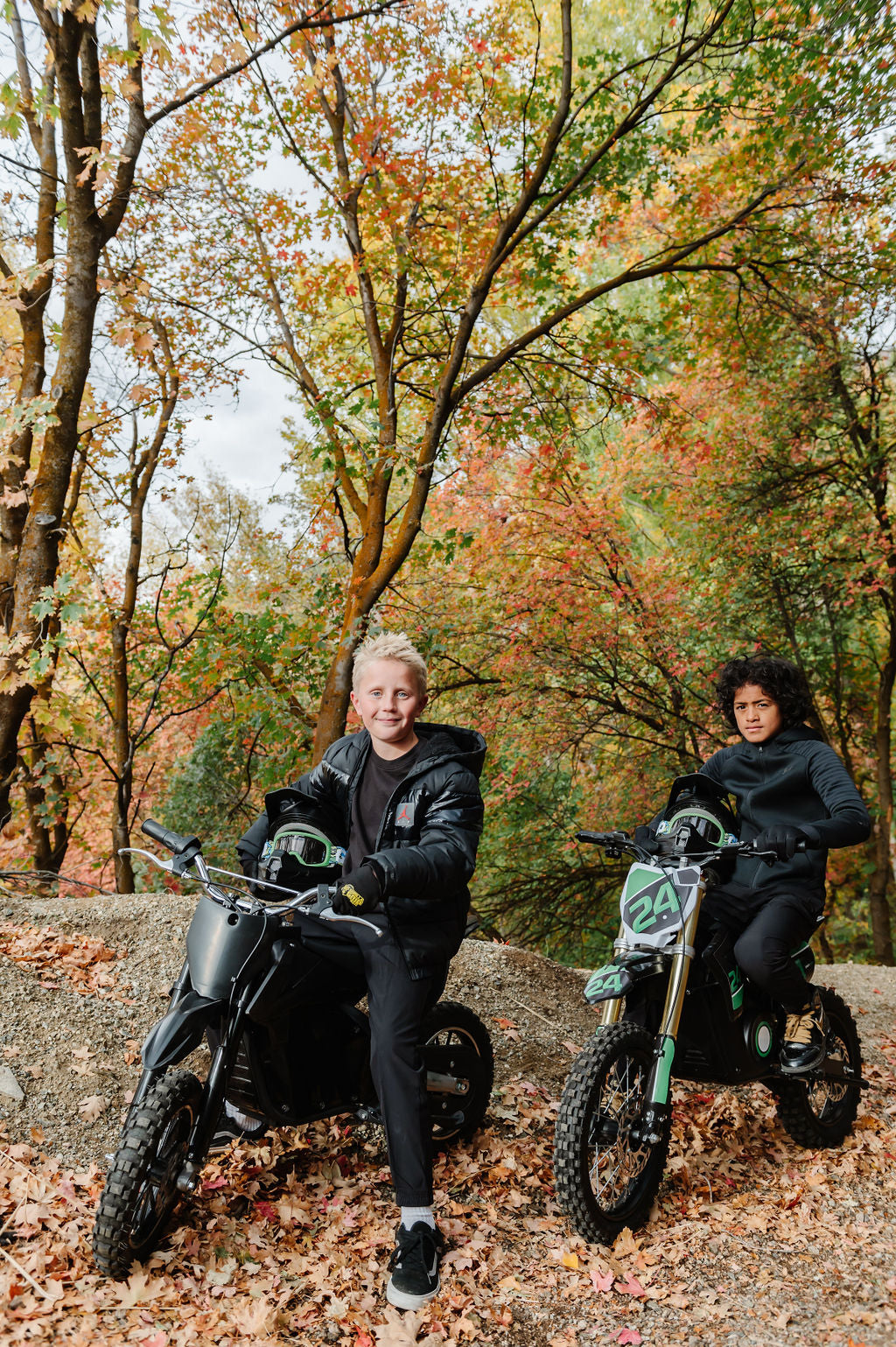 Two people sitting onDrift Hero USA 1600W Electric Dirt Bike in a forest with autumn foliage
