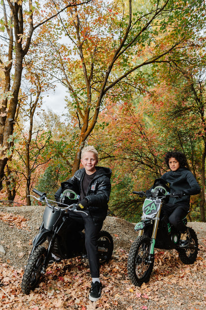 Two people sitting onDrift Hero USA 1600W Electric Dirt Bike in a forest with autumn foliage
