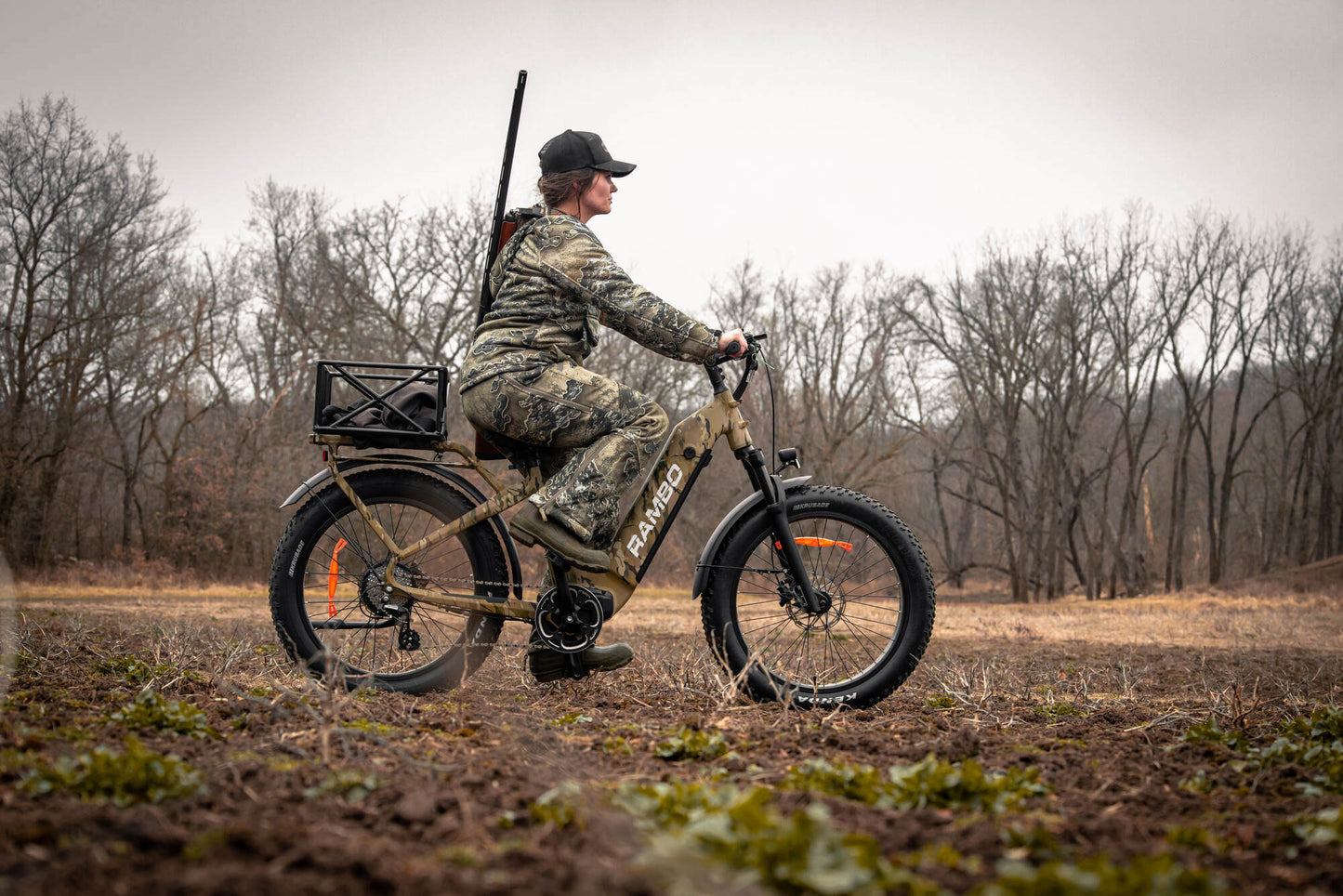 Person riding a camouflage Rambo Bikes SAVAGE 2.0 Electric Bike in a field with bare trees in the background