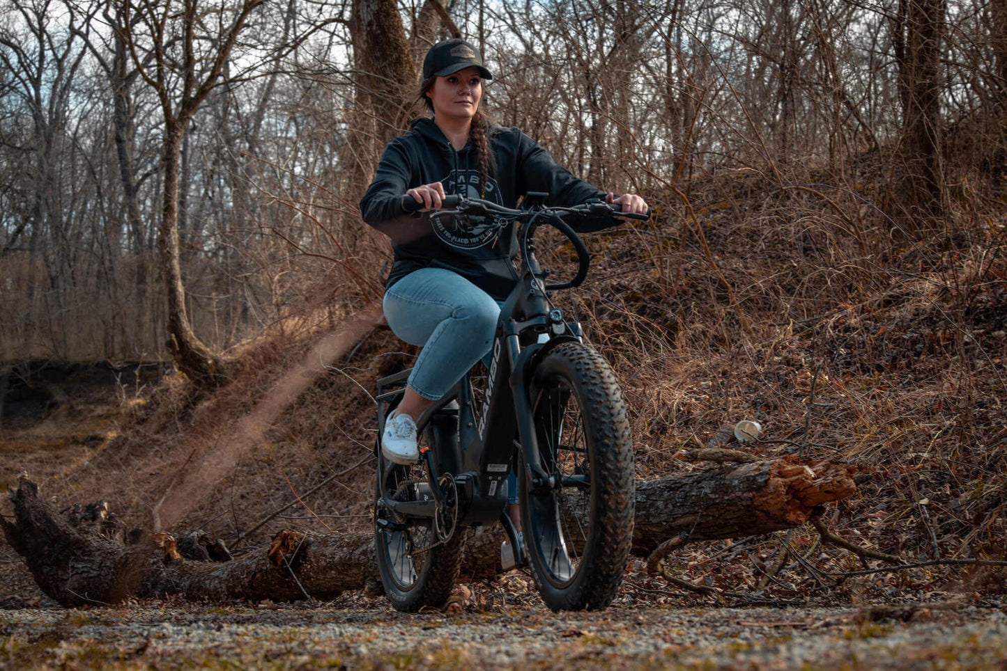 Person riding a Rambo Bikes SAVAGE 2.0 Electric Bike through a forest with bare trees and a fallen log.